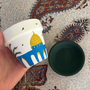 Puede incluir: Maceta de terracota pintada a mano con un diseño azul y blanco que presenta un edificio con cúpula amarilla. La maceta está sobre un platillo verde.