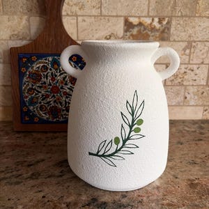 May include: White ceramic vase with two handles and a textured surface. The vase features a green olive branch design. A wooden cutting board with a decorative tile is in the background.