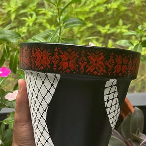Op de afbeelding: Zwart geschilderde bloempot met een rood en zwart bloemmotief rond de rand. De pot heeft een wit visnetpatroon op een zwarte achtergrond. De pot wordt in een hand gehouden, met groene bladeren op de achtergrond.