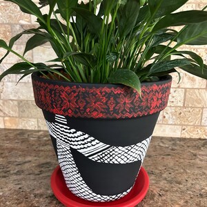May include: A black terracotta planter with a red and white patterned band around the top. The planter has a white snake design painted on the front and is sitting on a red saucer.