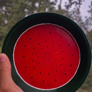 May include: A round, hand-painted decorative plate with a watermelon design. The plate features a bright red center with black seed details, a white border, and a dark green rim. The plate is held up in front of a window.