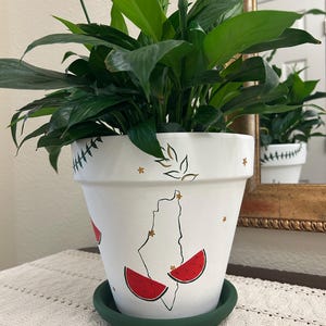 May include: A white ceramic plant pot with a green saucer, featuring a hand-painted design of a country outline and watermelon slices. The pot is filled with a lush green plant. The pot is sitting on a table with a white lace runner.