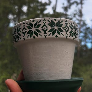May include: A white ceramic flower pot with a green snowflake pattern around the rim, sitting on a dark green saucer. The pot has a textured surface and is designed for planting flowers or herbs.