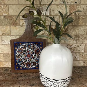 May include: A white ceramic vase with a black geometric pattern at the base, holding olive branches. A wooden cutting board with a decorative tile featuring a floral design is to the left.