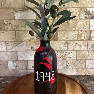 Op de afbeelding: Een zwarte vaas met rode verfspatten en het nummer "1948" in het wit. De vaas bevat olijftakken met groene bladeren en olijven. De vaas staat op een houten dienblad.