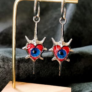 May include: A pair of silver earrings featuring a heart-shaped design with a red enamel finish. Each heart has a blue eye in the center and is surrounded by silver spikes. The earrings hang from silver hoops.