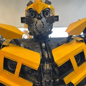 May include: A yellow and black robot made of scrap metal. The robot has a large head with a face mask and large eyes. The robot's body is made of various metal parts, including gears, bolts, and pipes.