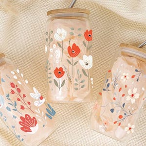 May include: Three glass drinking jars with floral designs. Each jar has a bamboo lid and a stainless steel straw. The floral patterns include red, white, and blue flowers and green leaves. The jars are filled with tissue paper.