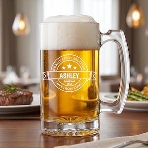 May include: A clear glass beer mug filled with golden beer and white foam. The front features a white circular graphic with the text "WORLD'S BEST RELATION", "ASHLEY", "ESTABLISHED 1957", and "LOVE FROM EMMA" centered within a banner design.