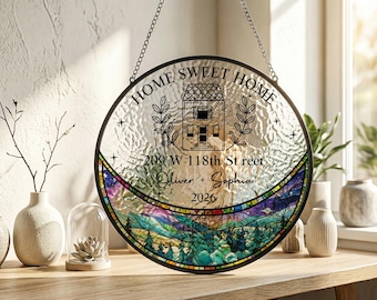 Puede incluir: Un atrapasoles circular de vidrieras con un centro transparente texturizado con el texto HOME SWEET HOME, una ilustración de casa, 209 W 118th St reet, Oliver + Sophia y 2026. El arco inferior muestra un paisaje colorido de montaña y bosque.