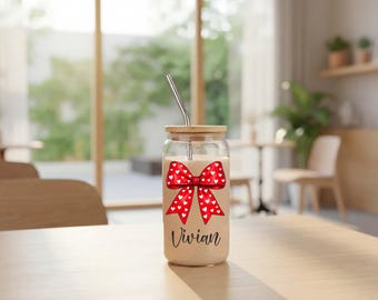 May include: A clear glass tumbler with a bamboo lid and a metal straw, filled with a beige liquid. The glass features a large red bow graphic with white heart patterns and the name Vivian printed in black script lettering below the bow.