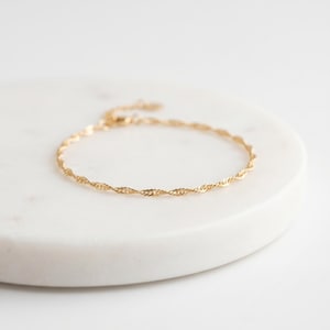 Op de afbeelding: Een delicate goudkleurige armband met een gedraaid touwontwerp. De armband wordt gepresenteerd op een wit marmeren oppervlak. Het sieraad heeft een verstelbare ketting en sluiting.