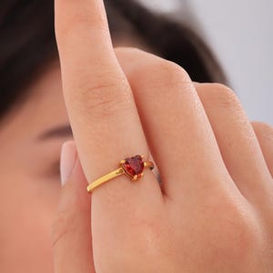 Peut inclure: Une bague en or ornée d'une pierre précieuse rouge foncé en forme de cœur. La bague a un fin anneau et une monture simple. La pierre en forme de cœur est le point central de la bague.