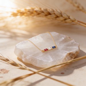 May include: A gold chain necklace featuring a horizontal bar pendant set with three rectangular baguette-cut gemstones in a row, consisting of a red stone, a clear white stone, and a deep blue stone, connected by a delicate gold link chain.