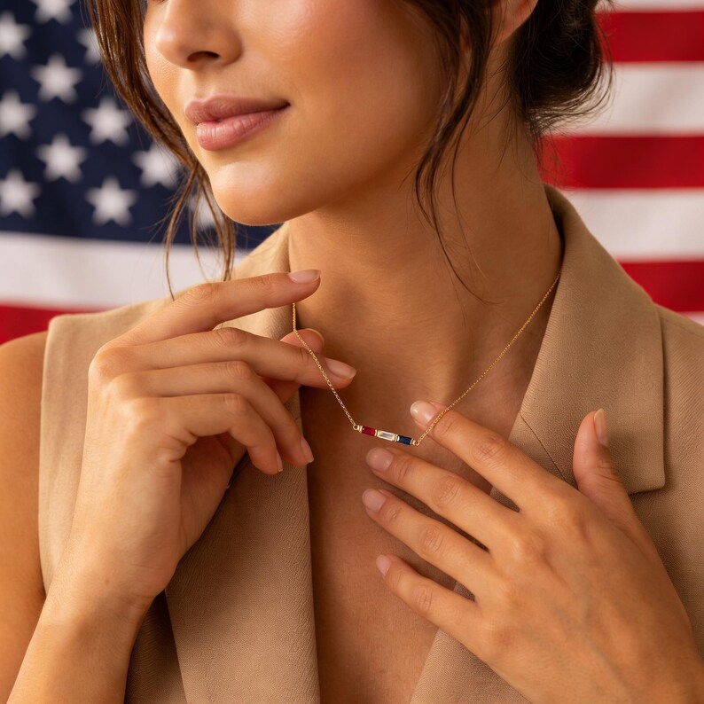 May include: A delicate gold chain necklace featuring a horizontal bar pendant set with three rectangular baguette-cut gemstones in a row, consisting of a red stone on the left, a clear white stone in the center, and a blue stone on the right side.