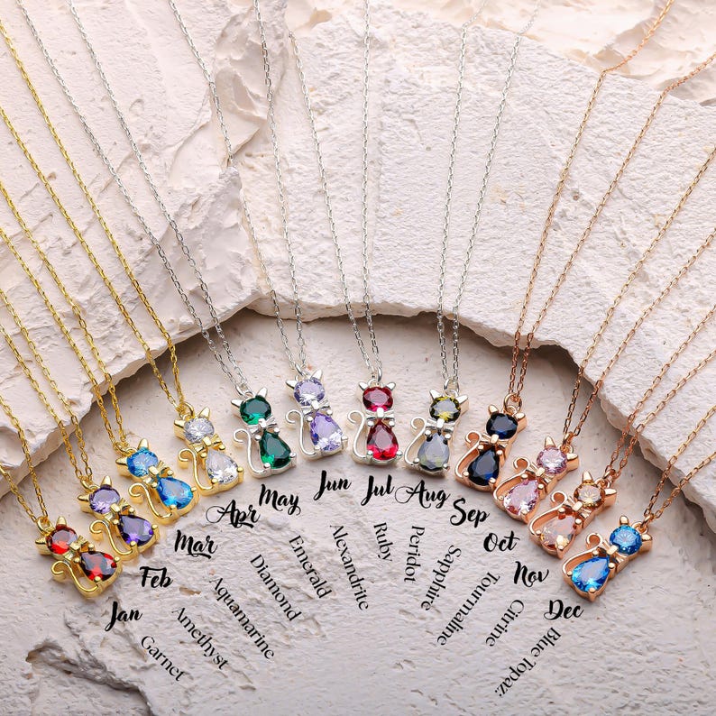 May include: A collection of cat-shaped pendant necklaces with various gemstone colors, including ruby, sapphire, and emerald. Each necklace has a delicate chain and is labeled with a month and gemstone name. The text includes "Jan" to "Dec".
