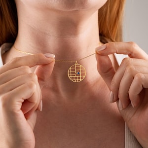 May include: A gold necklace with a circular pendant, a grid pattern, and a small blue gemstone. The necklace is held up, displaying the fine chain and pendant. The pendant's diameter is approximately 2 cm.