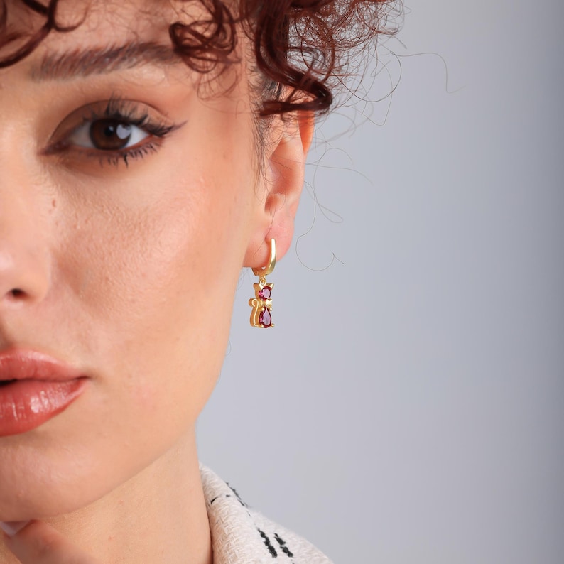 May include: Gold-colored hoop earrings with a cat pendant. The cat pendant features a red gemstone body and head. The earrings are worn on a person's ear, with a neutral background.