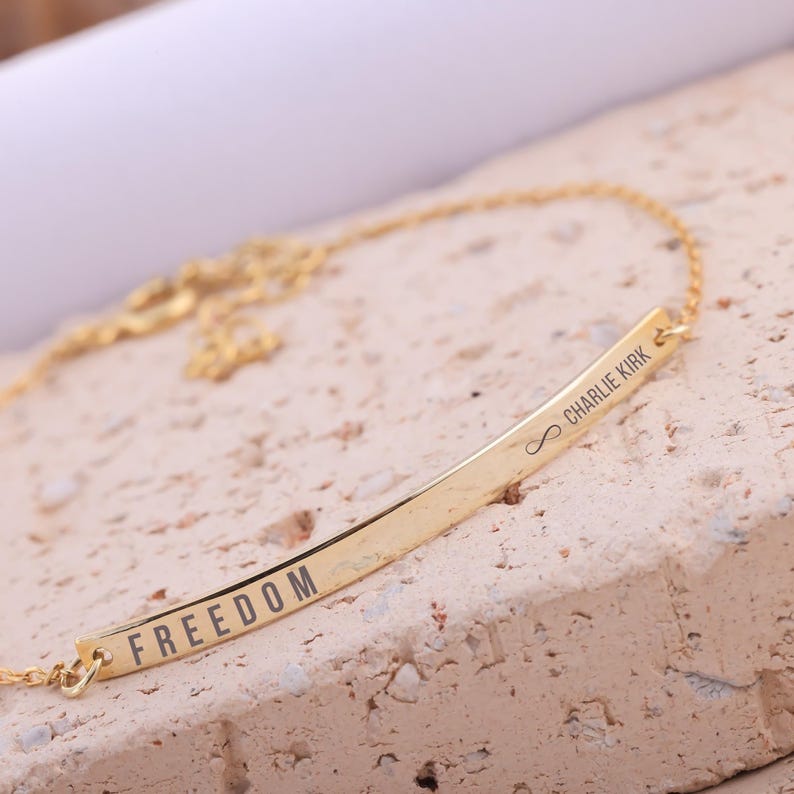 May include: A gold-colored necklace with a bar pendant. The word "FREEDOM" is engraved on the bar. The name "Charlie Kirk" and an infinity symbol are engraved on the other side of the bar. The necklace is displayed on a textured, light-colored surface.