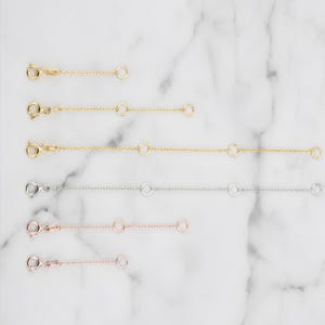 Peut inclure: Plusieurs rallonges de colliers en or, argent et or rose. Chaque rallonge est dotée d'un fermoir mousqueton à une extrémité et d'un petit anneau à l'autre, avec des anneaux supplémentaires pour le réglage de la longueur. Présentées sur une surface en marbre blanc.