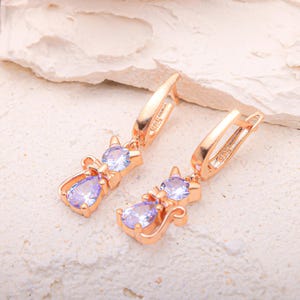 May include: Rose gold cat-shaped earrings with clear and light purple gemstones. The earrings feature a cat silhouette with a round gemstone head and a pear-shaped gemstone body. The earrings have a lever back closure.