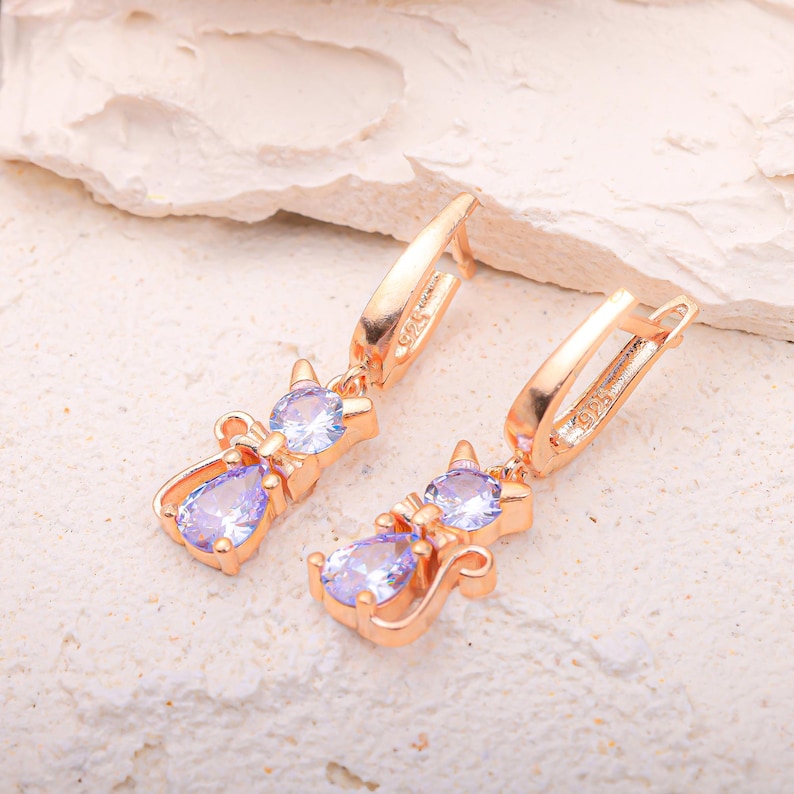 May include: Rose gold cat-shaped earrings with clear and light purple gemstones. The earrings feature a cat silhouette with a round gemstone head and a pear-shaped gemstone body. The earrings have a lever back closure.