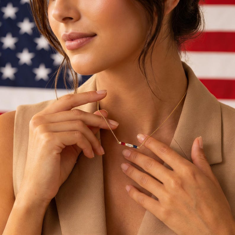 May include: A delicate gold chain necklace featuring a horizontal bar pendant set with three rectangular gemstones: a red stone on the left, a clear white stone in the center, and a dark blue stone on the right, resting against a tan top.