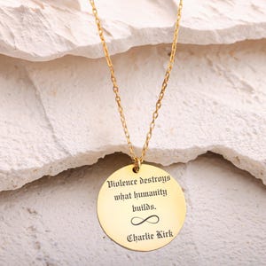 May include: A gold-colored necklace with a circular pendant. The pendant has the inscription "Violence destroys what humanity builds." below an infinity symbol, and "Charlie Kirk" at the bottom. The necklace is displayed against a textured, off-white background.