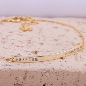 May include: A gold-colored bracelet with a curved bar pendant. The word "FREEDOM" is engraved on the bar. The bracelet also features the name "Charlie Kirk" and an infinity symbol. The bracelet is on a textured surface.