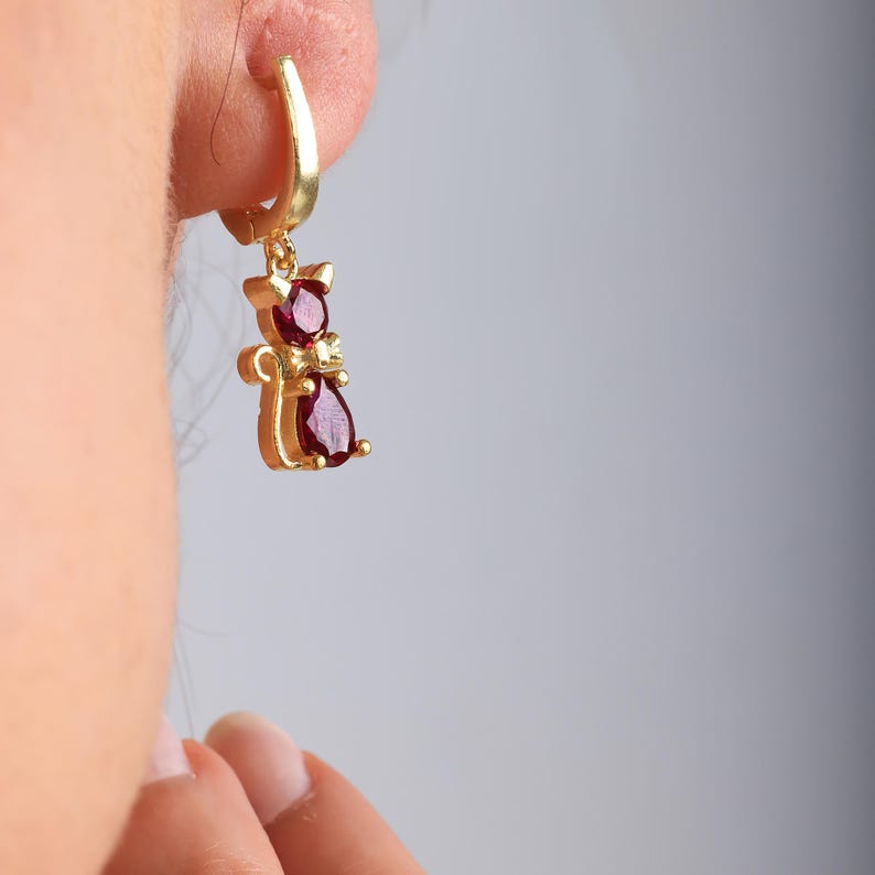May include: Gold-colored cat-shaped earring with a red gemstone. The earring is a small, dangling charm with a polished finish. The cat design features a round gemstone for the head and a teardrop-shaped gemstone for the body.