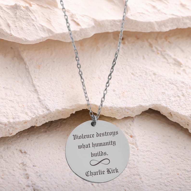 May include: Silver-toned necklace with a circular pendant. The pendant features the quote "Violence destroys what humanity builds." below an infinity symbol, and the name "Charlie Kirk". The necklace hangs against a textured, off-white background.