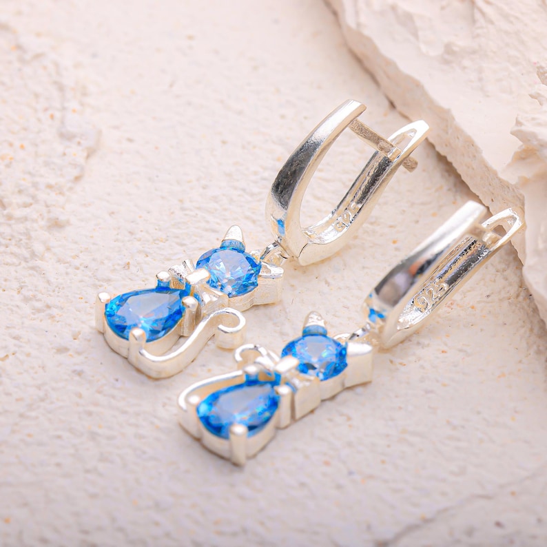 May include: A pair of silver cat-shaped earrings with blue gemstone accents. Each earring features a cat silhouette with a round blue gem at the head and a teardrop-shaped blue gem dangling below. The earrings have a lever-back closure.