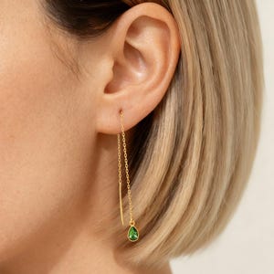 May include: A gold-colored earring with a delicate chain and a small, teardrop-shaped green gemstone. The earring is shown dangling from an ear, highlighting its elegant design and color.