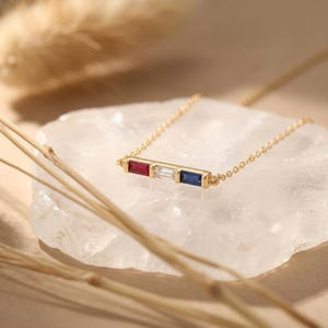 May include: A gold chain necklace featuring a horizontal bar pendant set with three rectangular baguette-cut gemstones: a red ruby on the left, a clear white diamond in the center, and a deep blue sapphire on the right side of the setting.