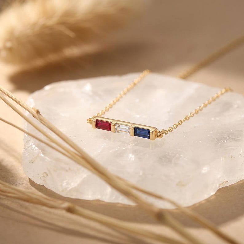 May include: A gold chain necklace featuring a horizontal bar pendant set with three rectangular baguette-cut gemstones: a red ruby on the left, a clear white diamond in the center, and a deep blue sapphire on the right side of the setting.