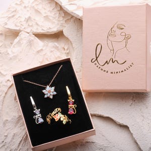 May include: A jewelry set in a light pink box with the text Daphne Minimalist. The set includes a flower pendant necklace with white stones, two cat-shaped earrings with red and iridescent stones, and a gold ring with the word Trinity and floral accents.