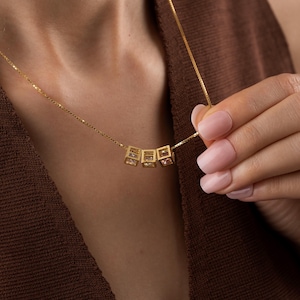 May include: A gold-colored necklace featuring a delicate chain and three square pendants. Each pendant has a cubic design with a gemstone inside. The necklace is displayed against a brown background.