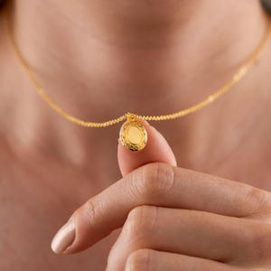 Może przedstawiać: Złoty naszyjnik z małą, owalną zawieszką w kształcie medalionu. Zawieszka ma ozdobny, grawerowany wzór na krawędziach. Naszyjnik jest trzymany przez palec osoby, ukazując delikatny łańcuszek i szczegóły zawieszki.