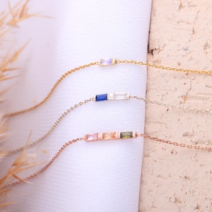 May include: Three delicate gold, silver, and rose gold chain bracelets with colorful gemstone accents. Each bracelet features a row of small, rectangular gemstones in different colors, including pink, blue, and green.