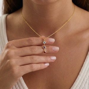 May include: A gold-coloured necklace featuring three gemstones in light blue, purple, and red. The gemstones are arranged vertically from the chain. The necklace is being held up by a person.