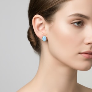 Könnte beinhalten: Nahaufnahme eines Ohrs mit einem ovalen, hellblauen Opal-Ohrstecker in Goldfassung. Die Ohrringe stehen im Mittelpunkt, vor einem neutralen Hintergrund. Der Opal hat einen subtilen Schimmer.