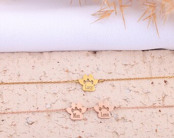 May include: Three delicate gold and rose gold bracelets. Each features a paw print charm with a name engraved: "Lucy", "Max", and "Luna". The bracelets are displayed on a textured surface with dried plants in the background.