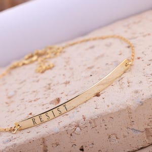 May include: A gold-colored necklace with a bar pendant. The pendant is engraved with the word "RESIST" in capital letters. The necklace is displayed on a textured, light-colored surface.