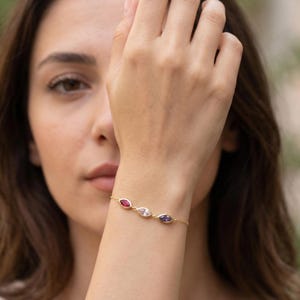 Könnte beinhalten: Goldenes Armband mit drei marquiseförmigen Edelsteinen in Rot, Klar und Lila. Das Armband ist zart und hat eine feine Goldkette. Die Edelsteine sind in einer Zargenfassung gefasst.