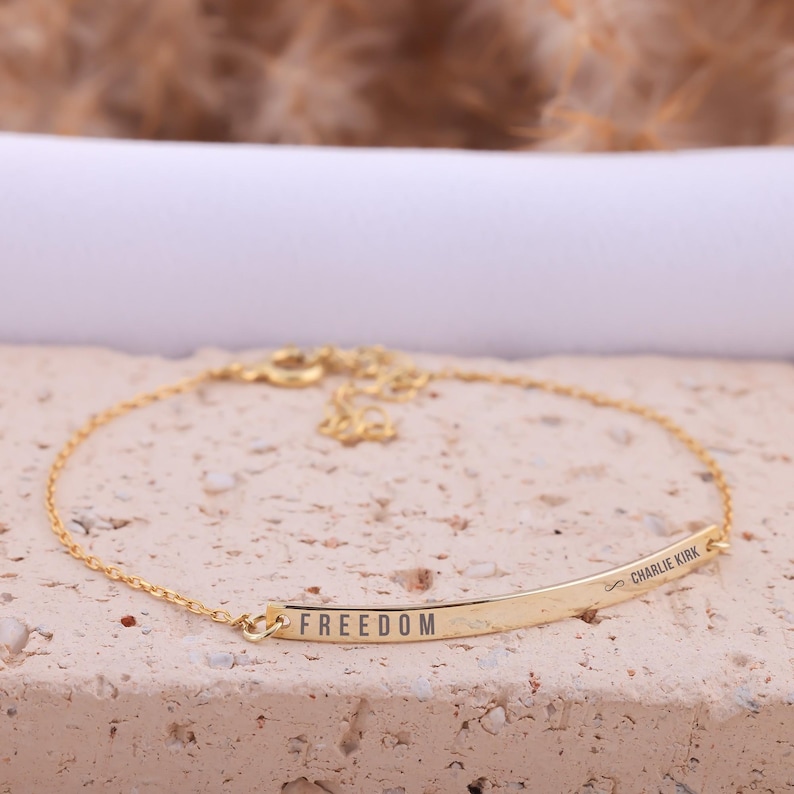 May include: A gold-colored bracelet with the word "FREEDOM" engraved on a curved bar. The bracelet has a delicate chain and a clasp. The name "Charlie Kirk" is also engraved on the bar. The bracelet is displayed on a textured surface.