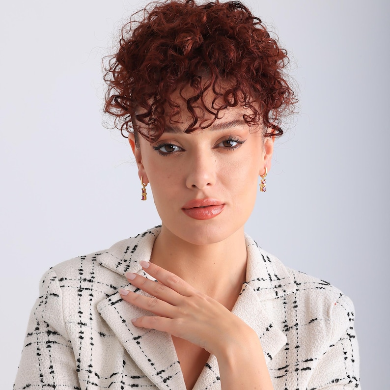 May include: Gold-colored hoop earrings with a small, rectangular charm. The earrings are worn by a person wearing a cream-colored blazer with a black plaid pattern. The person has curly auburn hair and is looking directly at the camera.