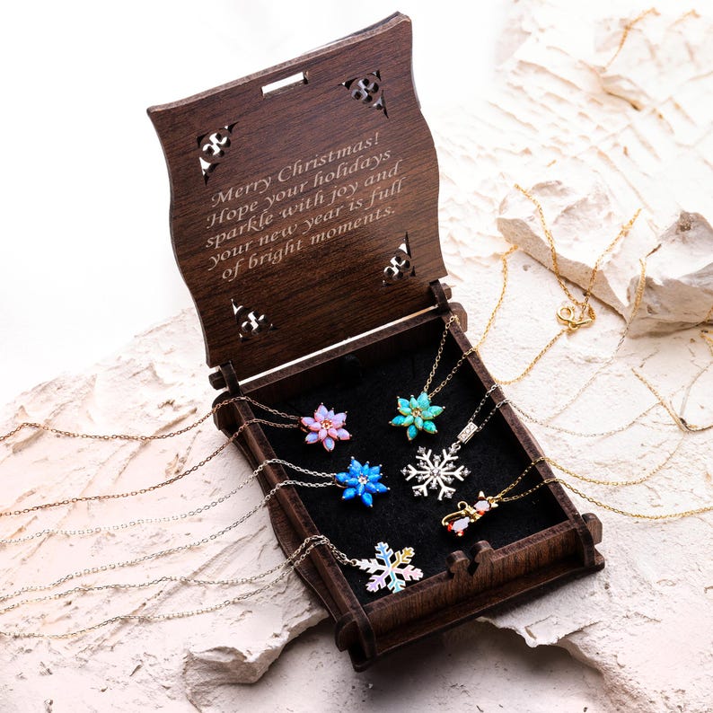 May include: A dark brown wooden box with a hinged lid featuring laser-cut corners and the engraved text Merry Christmas! Hope your holidays sparkle with joy and your new year is full of bright moments. Inside are six delicate necklaces with snowflake pendants.