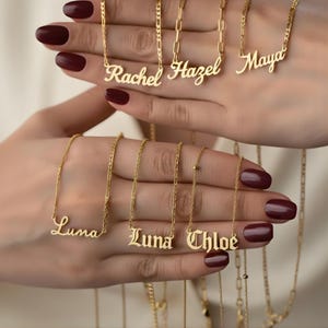 May include: Several gold-coloured name necklaces are displayed. The necklaces feature names in cursive and block lettering. The chains are delicate, with the names as the main focus. The background is a soft, neutral colour.
