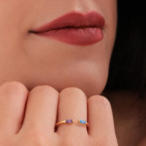 May include: A gold ring with an open design, featuring a purple and a blue rectangular gemstone. The ring is worn on a finger, with a person's lips and part of their face visible in the background.
