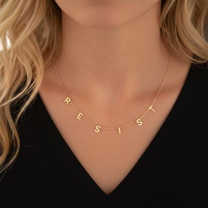 Peut inclure: Un délicat collier en or épelant le mot "RESIST" en lettres majuscules individuelles. Le collier est composé d'une fine chaîne et est porté sur un fond sombre. Les lettres sont espacées uniformément et semblent être en métal brillant.